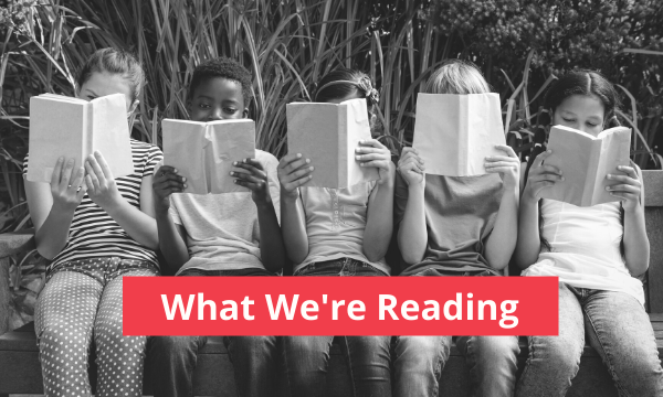 This image shows a black and white photo of a group of children sitting and holding books over their faces. The text says "What We're Reading"