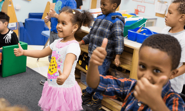 This image shows four young kids in a classroom setting giving a big thumbs up.