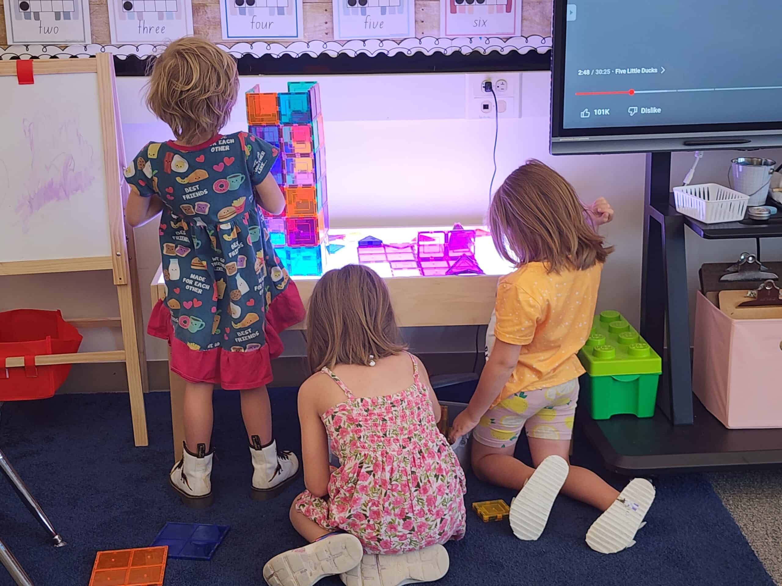 Three kids gather next to a light table during free play time.