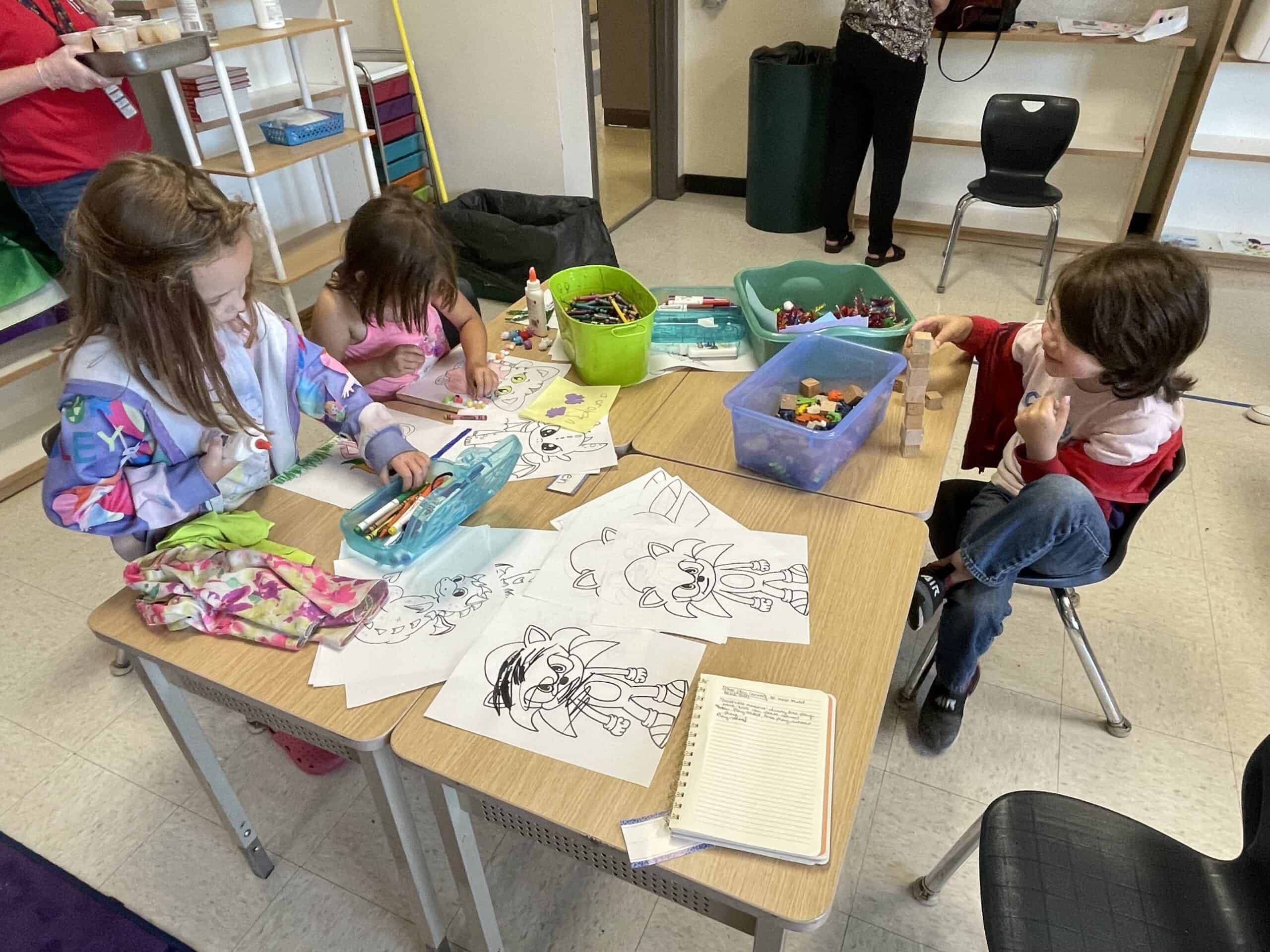 Three incoming kindergartners sit together at a table, covered with coloring pages. They are participaing in an activity together. One child is playing with what appears to be blocks, the other two children are coloring. 