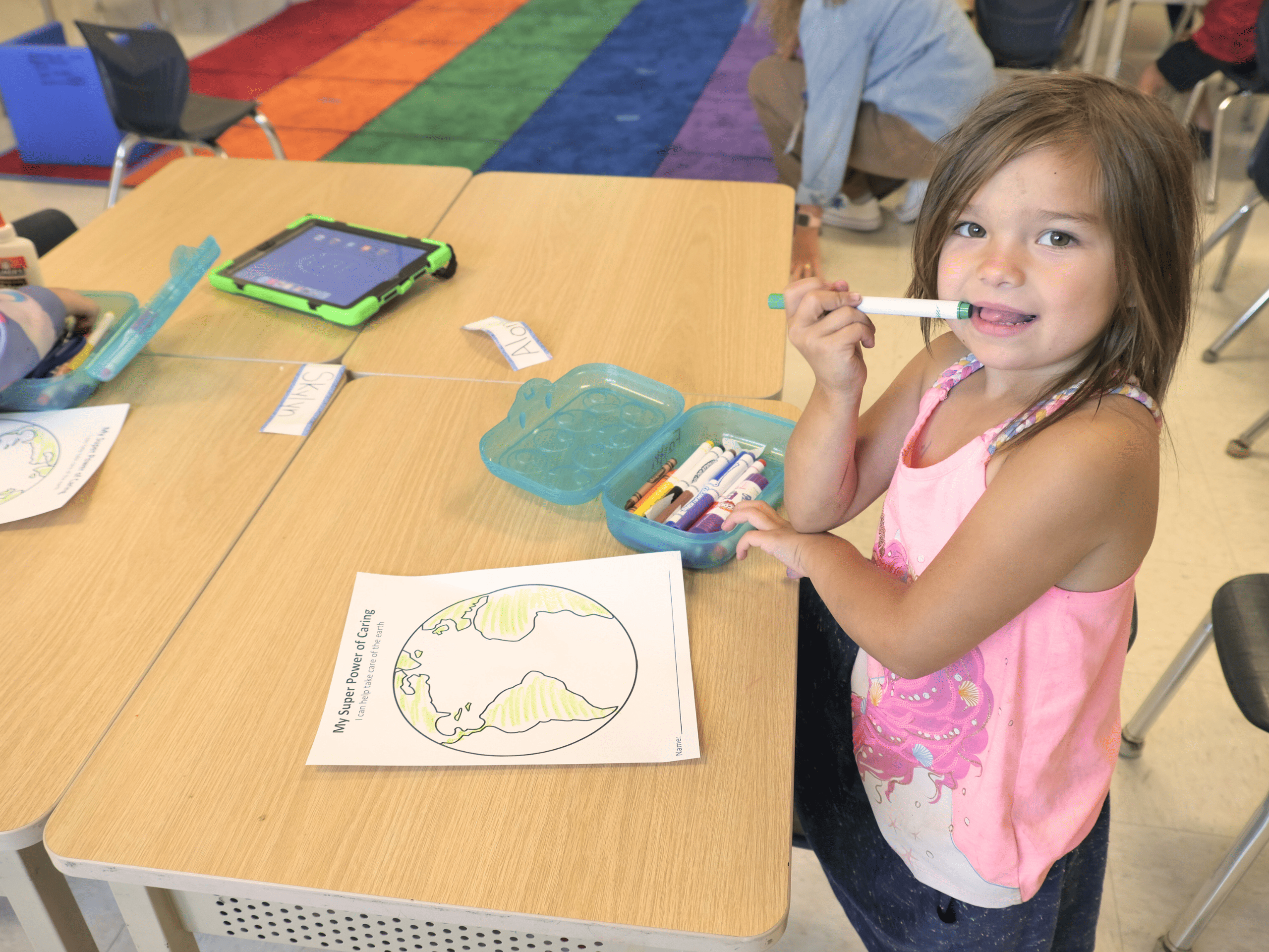 A young child sits at a desk with a marker in hand, near their mouth. A coloring sheet sits in front of the child, and a box of markers is nearby. 