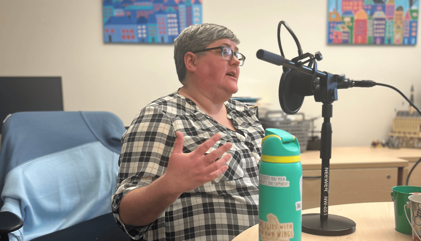 This image features our podcast guest Liane, who has short grey brown hair and is wearing a black and white checked shirt while sitting in front of the mic to record