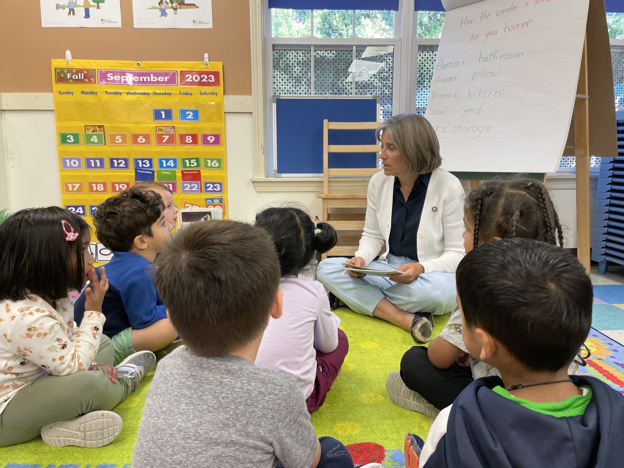 Sen. Lisa Reynolds, a pediatrician, sits on the floor in a classroom with a group of little children listening to her.