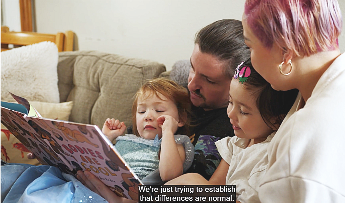 A family of four sits on a couch and reads a children's book. The photo is a still shot, so there's a snippet from the closed caption. It says: ”We’re just trying to establish that differences are normal.” Social justice is a major them of this film.